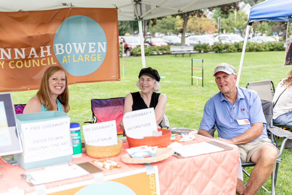 Photo of Hannah and two volunteers sitting under a tent at an outdoor event with campaign materials on a table in front of them
